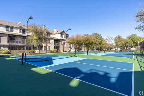 A tennis court with a blue surface and white lines is surrounded by apartment buildings.
