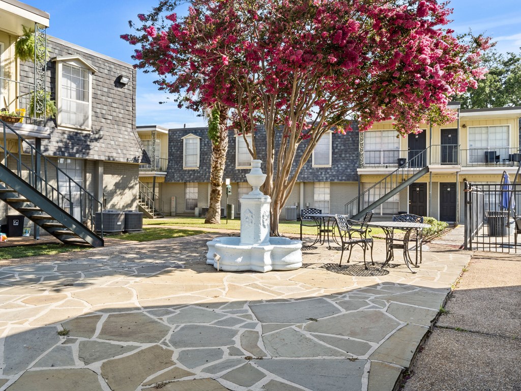 A tree with pink flowers is in the middle of a courtyard with a white pillar and a bench.
