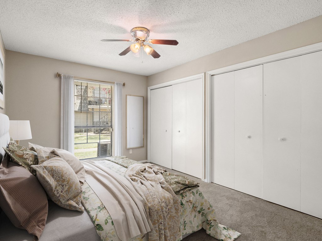 A bedroom with a bed, a ceiling fan, and a window with a view of a fence and trees.