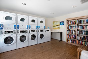 A laundry room with a row of washers and dryers, a bookshelf, and a table with a painting on the wall.