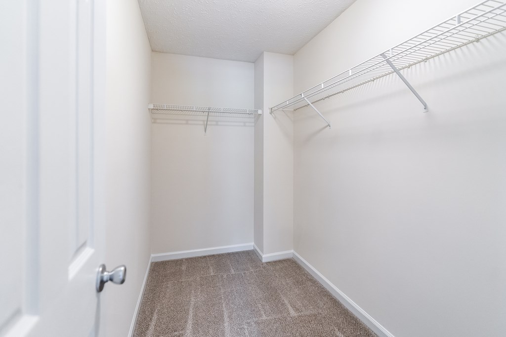 A white door is on the left of a hallway with a carpeted floor and a metal shelf on the wall.