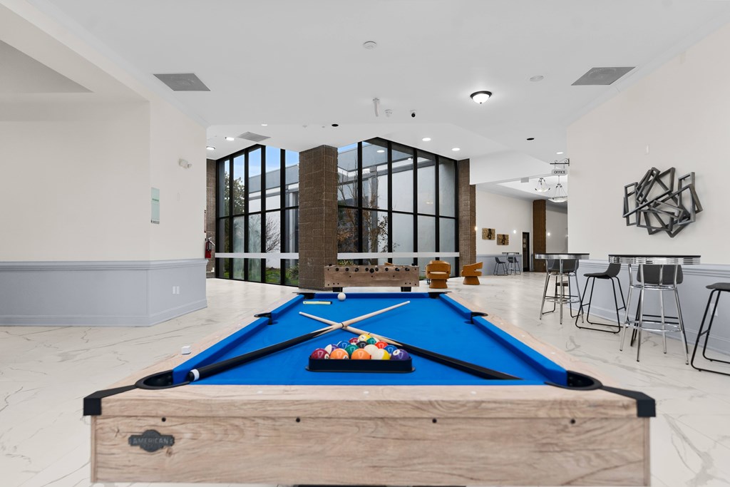 A pool table with a blue cloth and a wooden cue stick is in the foreground of a spacious room with a bar and a fireplace.