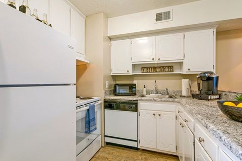 A kitchen with white appliances and cabinets with a sign that says "COFFEE-ANYONE?" on the wall.