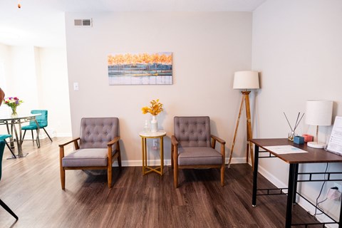 A living room with two grey chairs, a small table with a vase of flowers, and a painting on the wall.