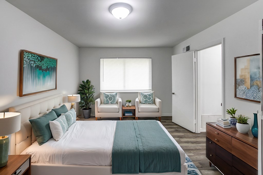 A bedroom with a bed, two chairs, a nightstand, and a painting on the wall.