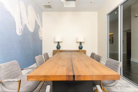 A long wooden table is surrounded by grey chairs in a room with a blue wall.
