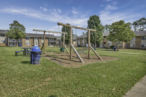 A playground with a swing set and a blue trash can in front of apartment buildings.