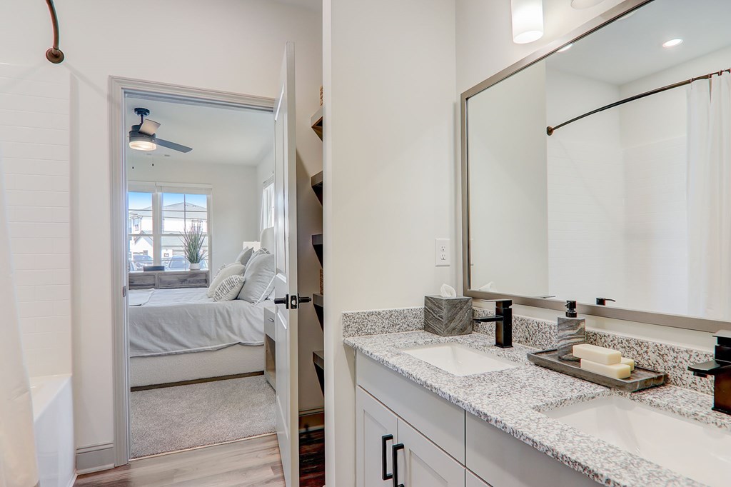 A bathroom with a mirror, sink, and a bedroom with a bed and a window.