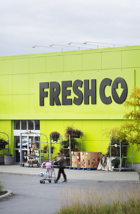 A person is pushing a shopping cart in front of a bright yellow FreshCo store.