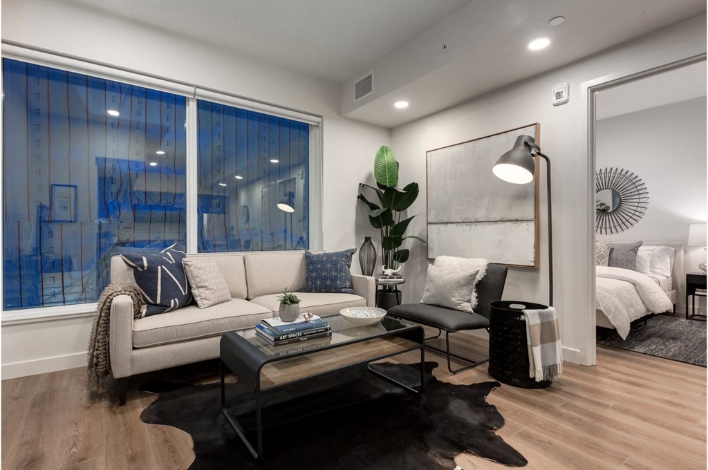 A modern living room with a large window, a sofa, a coffee table, and a bed in the background.