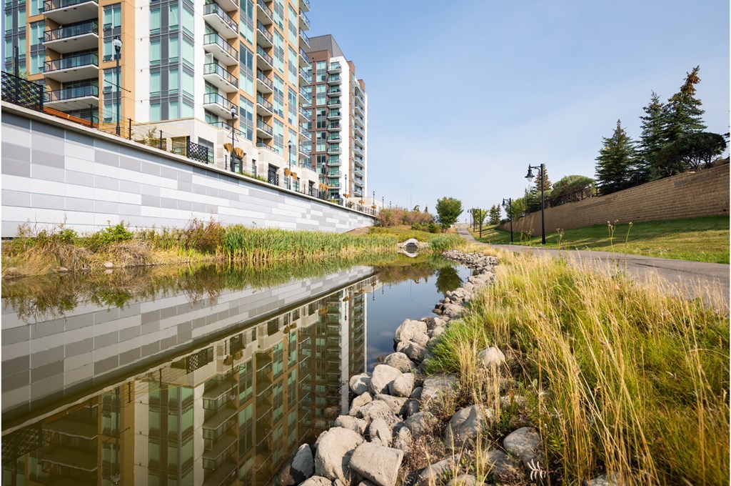 A river runs through a grassy area next to a concrete wall and apartment buildings.