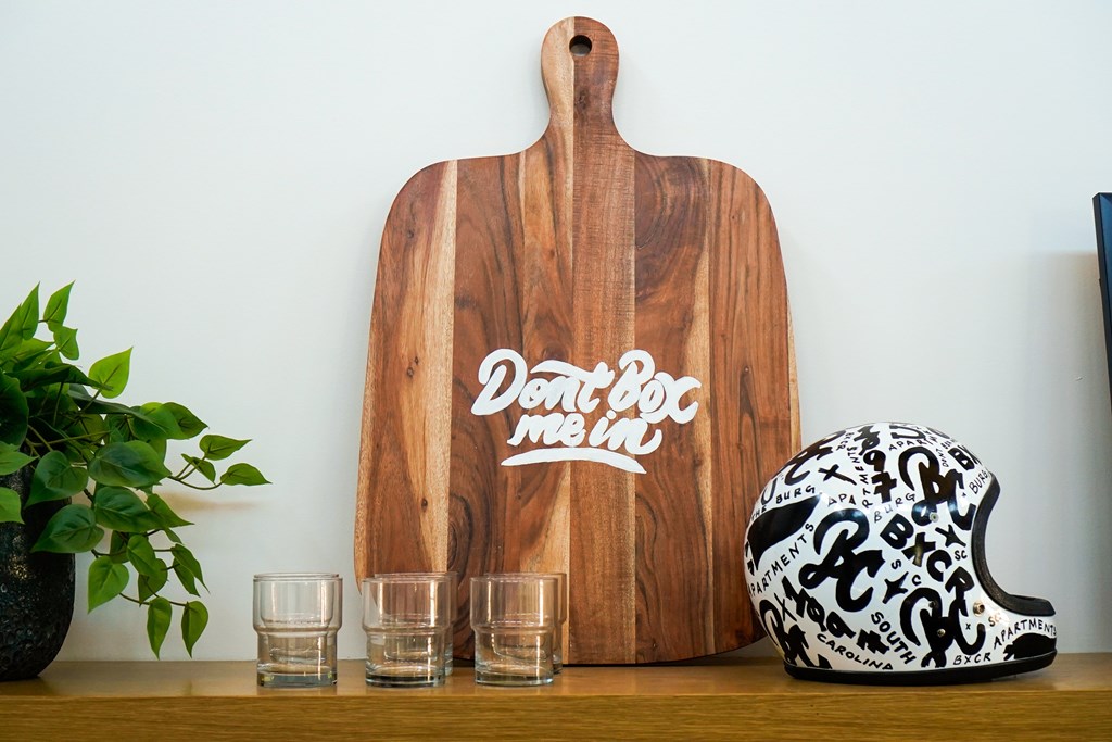 A wooden cutting board with "Don't Roc Me" written on it sits on a table next to two shot glasses and a helmet with black and white designs.