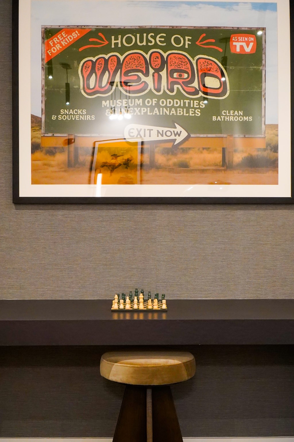 A framed poster of House of Weird is on the wall above a table with a chess set on it.