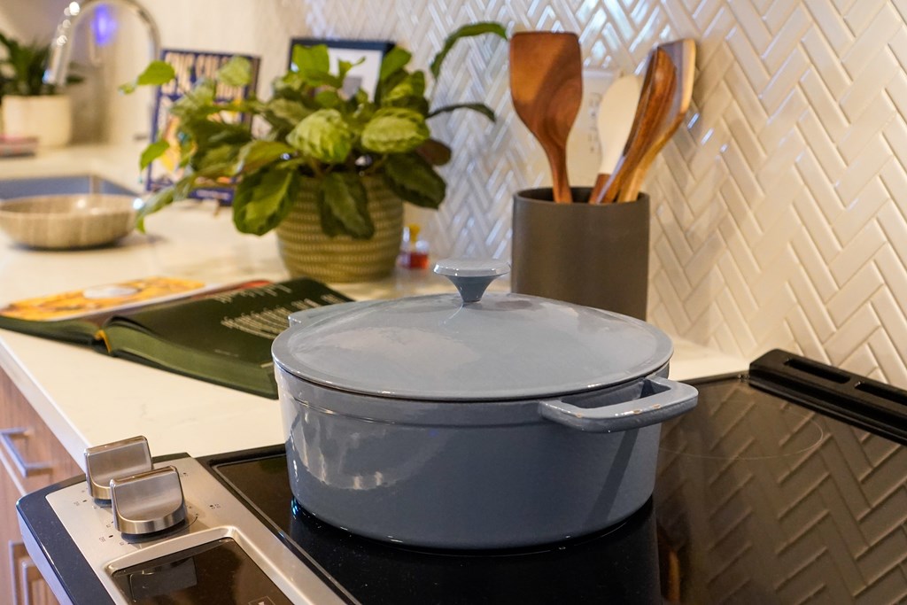 A pot sits on a stove with a plant in a vase and utensils in a holder in the background.