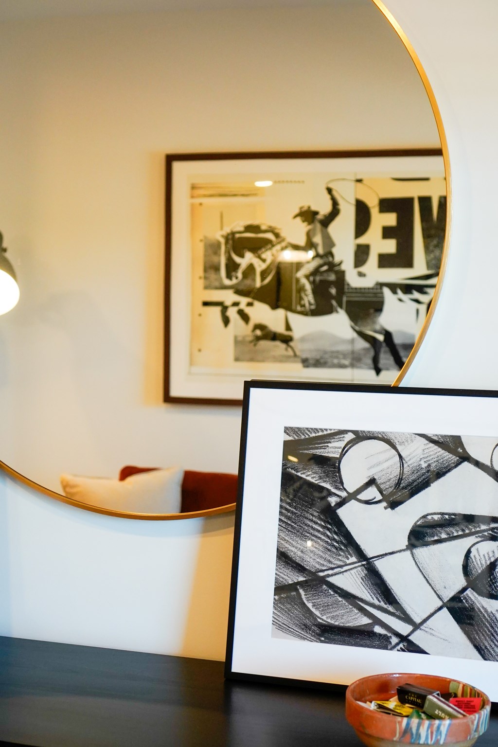A framed black and white abstract art piece sits on a table next to a round mirror.