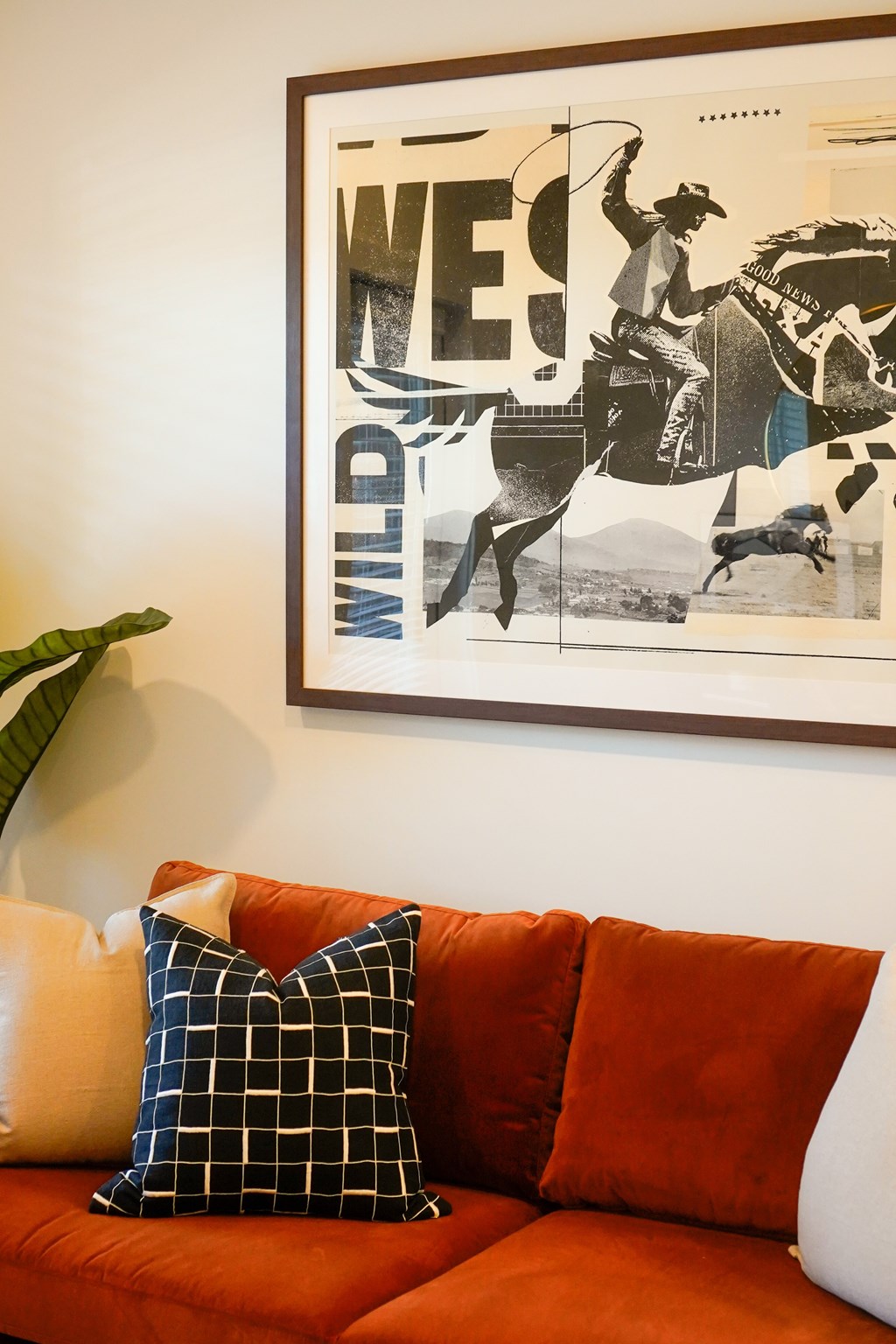 A framed poster with a man on a horse and the word "Wild" is on a wall above a red couch with two pillows.