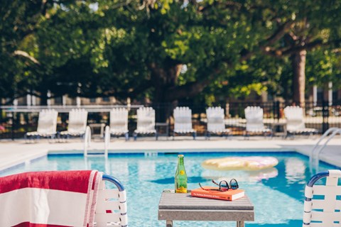 A table with a bottle, glasses and a book sits by a pool.