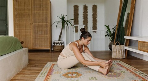 A woman is sitting on a colorful rug in a room with wooden floors and a white wall.