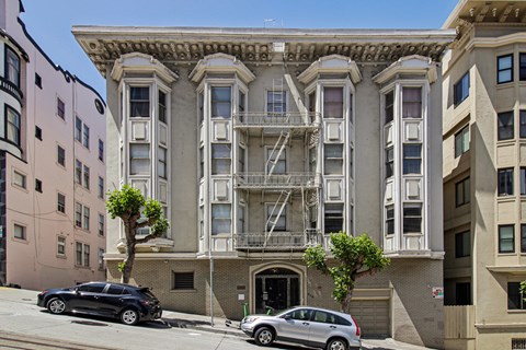 A black car is parked on the street in front of a tan building with a tree in front of it.