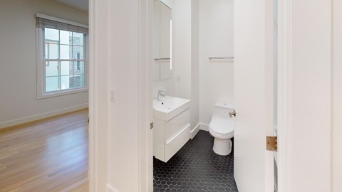 A small, modern bathroom with a toilet and sink on hexagonal black tiles. Adjacent is a bright room with wooden floors and a large window.
