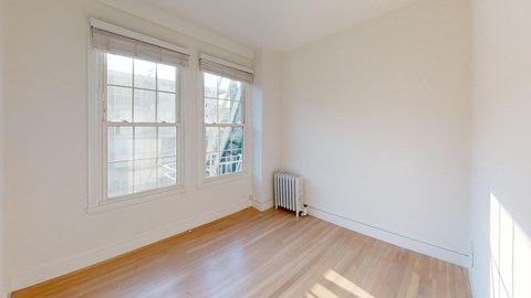Bright, empty room with hardwood floors, featuring two large windows on the left. A small radiator sits beneath one window.