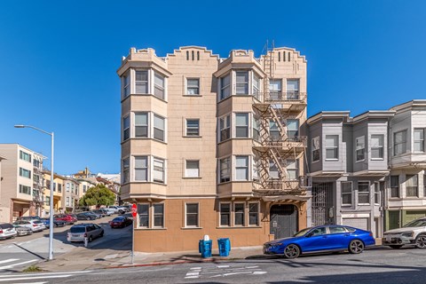 A blue car is parked in front of a tan building with a fire escape.