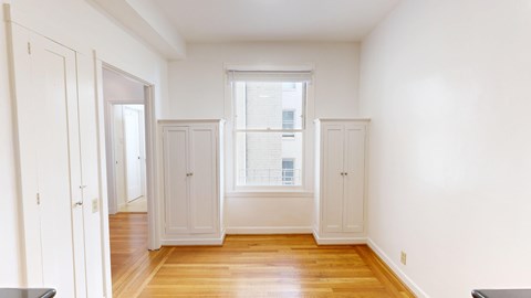 A room with white walls and wooden floors with a window and cupboards.