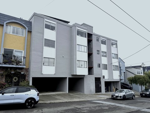 A white car is parked in front of a grey and yellow apartment building.