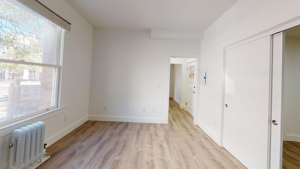 A room with white walls and wooden flooring with a window on the left side.