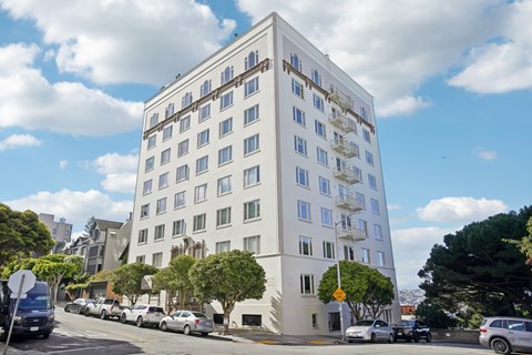 A white building with many windows is on a street with cars parked in front.