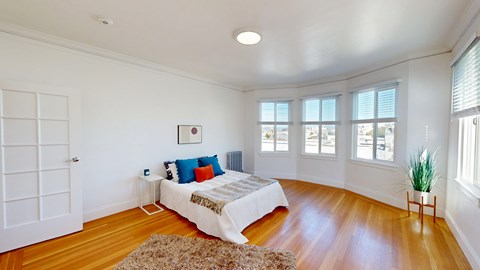 A bedroom with a bed, pillows, a rug, a small table, a potted plant, and a window.
