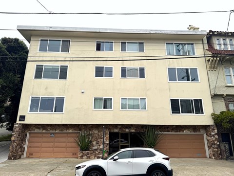 A white car is parked in front of a tan building.