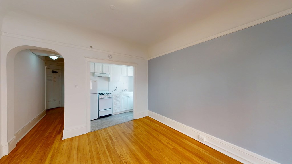 A room with wooden floors and a kitchen visible through the archway.