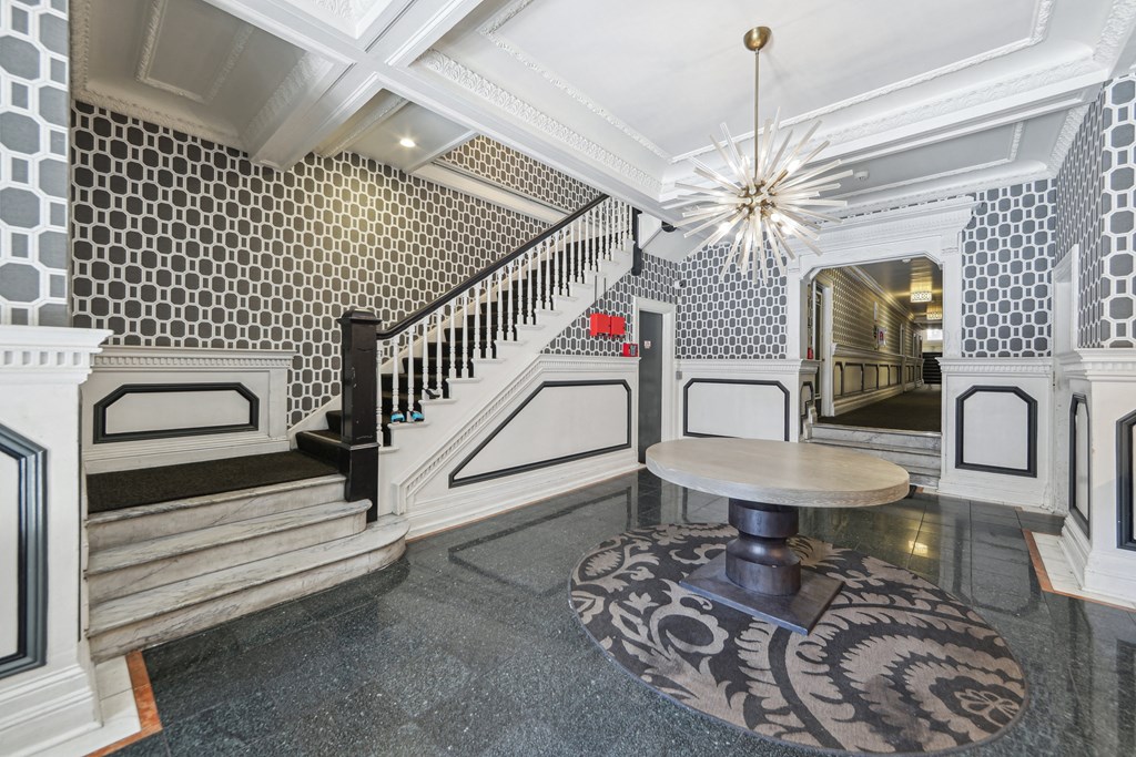 Elegant foyer with patterned wallpaper, a modern chandelier, unique round table, and decorative area rug, leading to a staircase