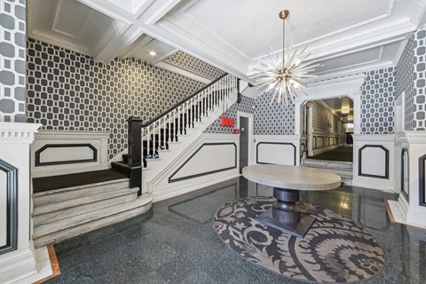 Elegant foyer with patterned wallpaper, a modern chandelier, unique round table, and decorative area rug, leading to a staircase