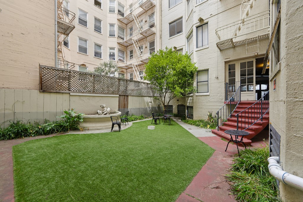 Courtyard with a wooden fence, with a small garden featuring a fountain and a statue, and a staircase that leads up the the entrance of the apartment building