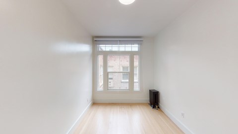 A small, empty room with wooden flooring, and a large window with blinds overlooking a brick building.
