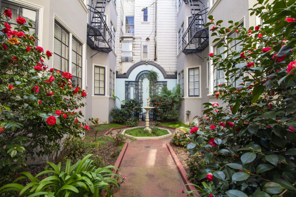 Charming apartment courtyard with lush greenery, red flowering plants, and an arched entryway leading to a fountain