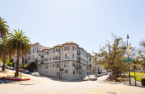 A street view of a residential area with apartment buildings, cars, and palm trees.