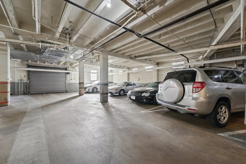 Underground parking garage with cars, marked spaces, lighting, and visible piping; closed shutter door