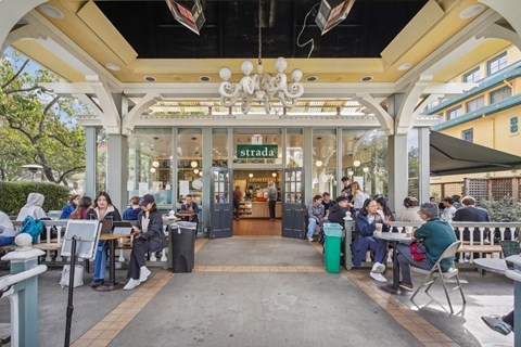 Bustling outdoor cafe with a large glass entrance framed by columns, filled with people seated at tables