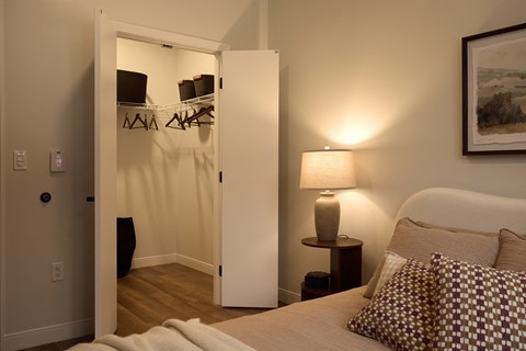A bedroom with a bed, a nightstand with a lamp, and a closet with clothes hanging.