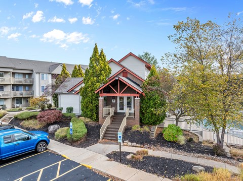 A small house with a red roof is surrounded by trees and a blue car is parked in the driveway.