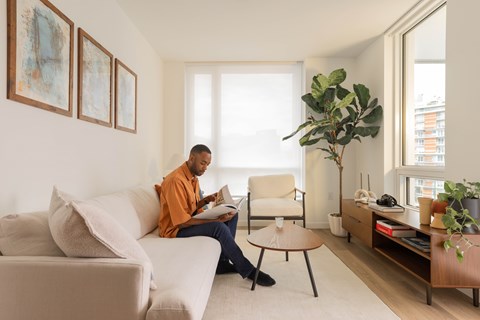 A man in an orange shirt is sitting on a beige couch reading a book.