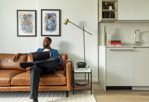 A man is sitting on a brown leather couch with a laptop on his lap.