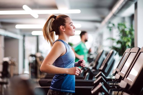 A woman in a blue top and grey shorts is running on a treadmill in a gym.
