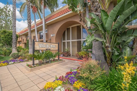 a building with a sign in front of a flower garden at Villa La Paz Apartments, Bellflower