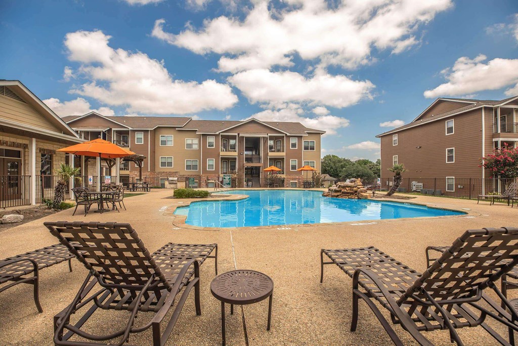 A pool surrounded by chairs and umbrellas in front of apartment buildings at The Villages at Lost Creek Apartments, San Antonio