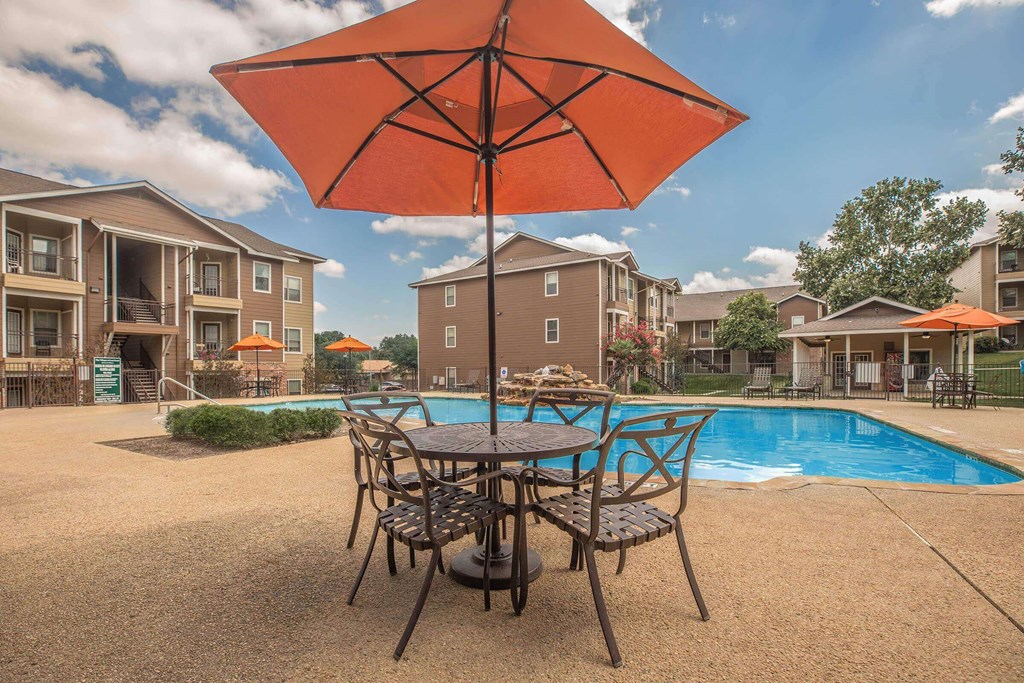 A patio with a table and chairs under an umbrella with a pool and apartment buildings in the background at The Villages at Lost Creek Apartments, Texas, 78247