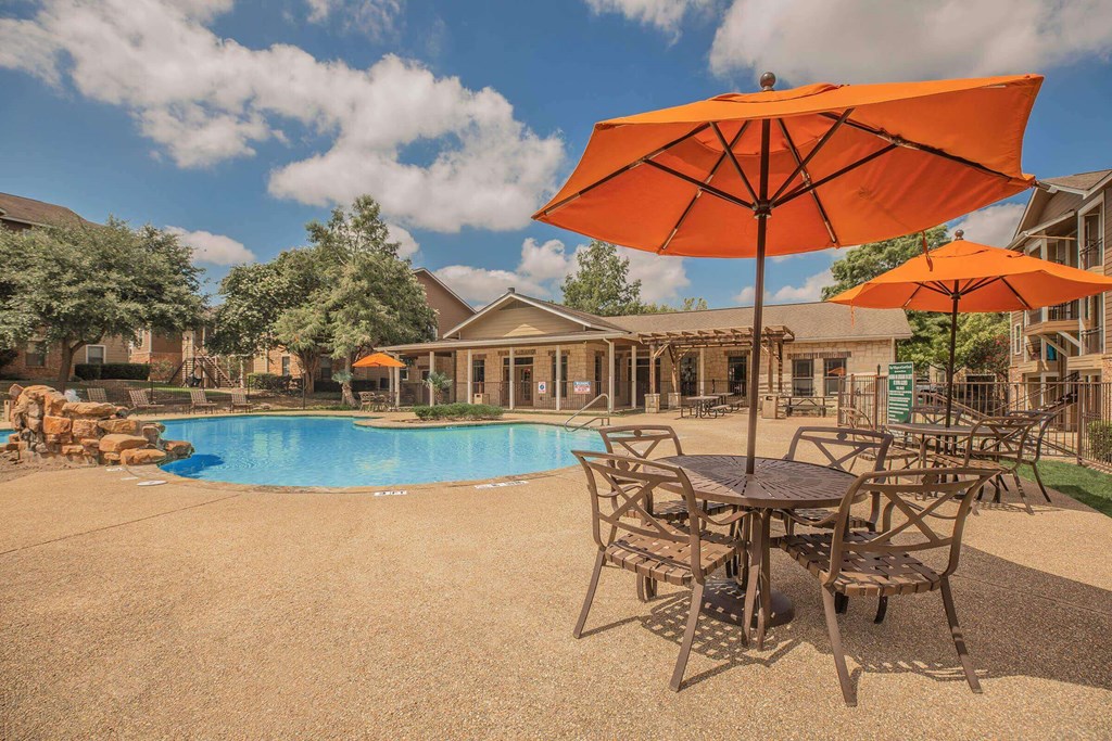 A large orange umbrella is in the foreground of a pool area. A large orange umbrella is in the foreground of a pool area at The Villages at Lost Creek Apartments, San Antonio, TX, 78247